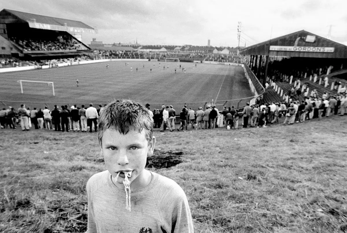 black and white photograph of young boy with toy skeleton in his mouth with soccer stadium in background