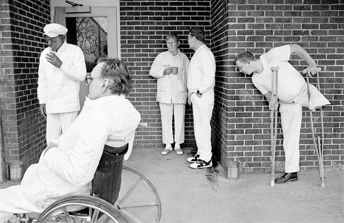 black and white photograph of people outside a care facility, man in wheelchair in foreground and man on crutches in background